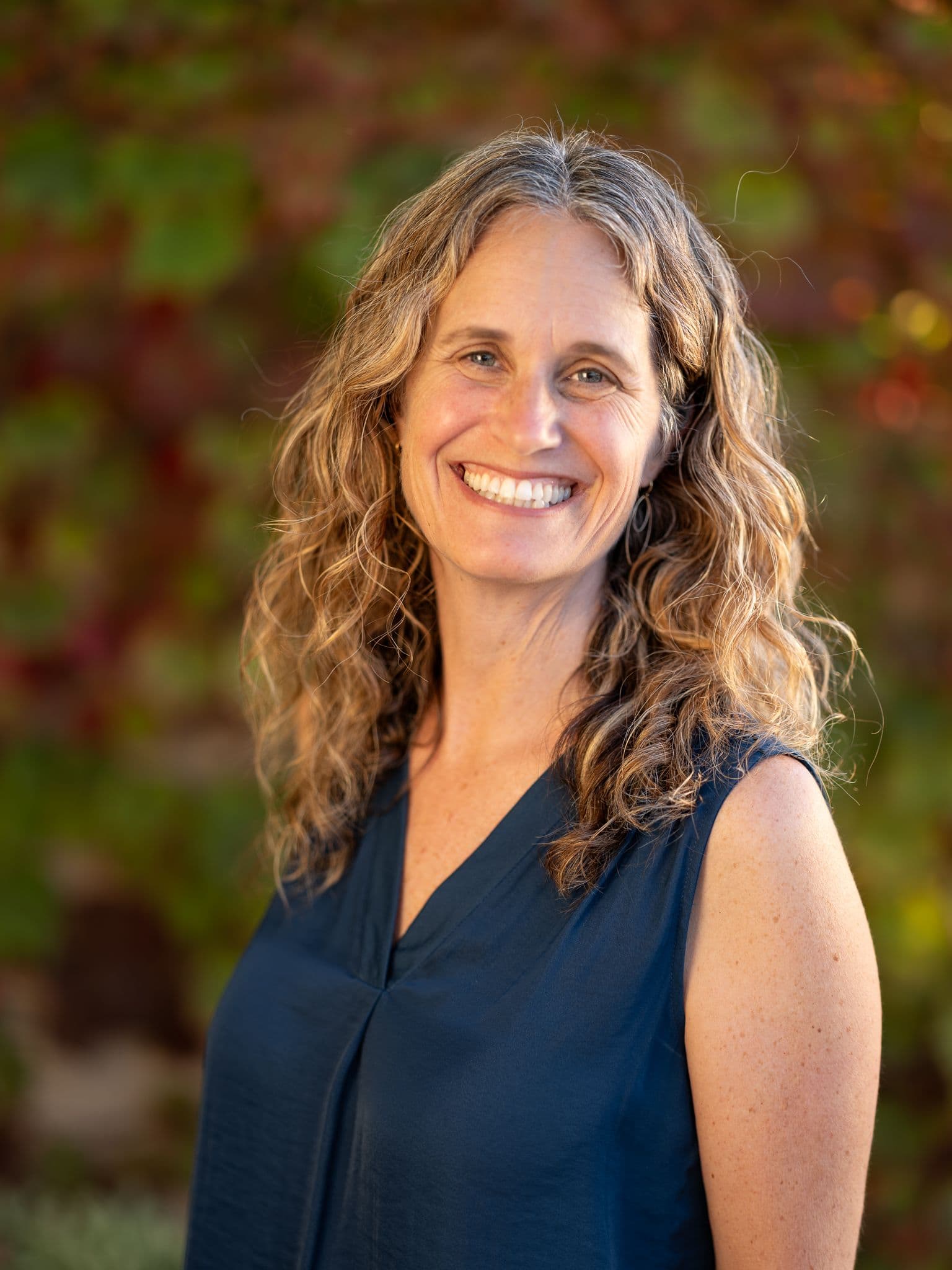 Robin Kaplan smiling outdoors in a navy blouse, with shoulder-length hair and a softly blurred background.